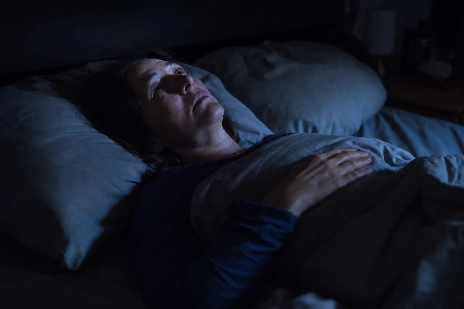 A woman in her fifties lying awake in a dark bedroom in the middle of the night, staring at the ceiling